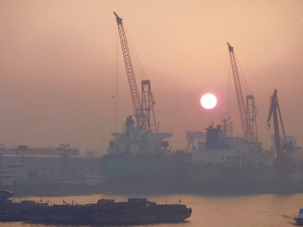 A blazing yellow sun and dusky orange sunset form a backdrop for several enormous cargo ships and towering cranes in the port of Shanghai.