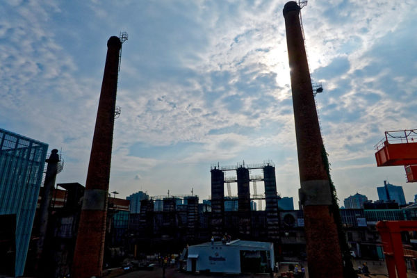 Two enormous red brick smokestacks tower over cranes and equipment at a construction site, rising high into a pale and cloudy sky.