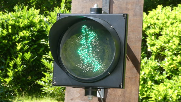 Against a backdrop of green trees and a brown wooden board, a metal traffic light displays a green outline of a running man who appears to be in motion.