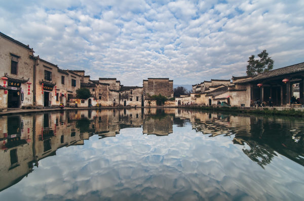 A small lake reflects the scenery that surrounds it, creating a top/bottom symmetry: two skies filled with puffy white clouds, and two villages of one- and two-story, mud-brick, tile-roofed houses adorned with red lanterns. Taken in Hongcun, a village in Anhui Province.