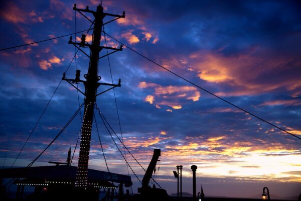 The silhouette of a ship’s mast adorned with tiny decorative lights towers against a dramatic and stormy sunset. 