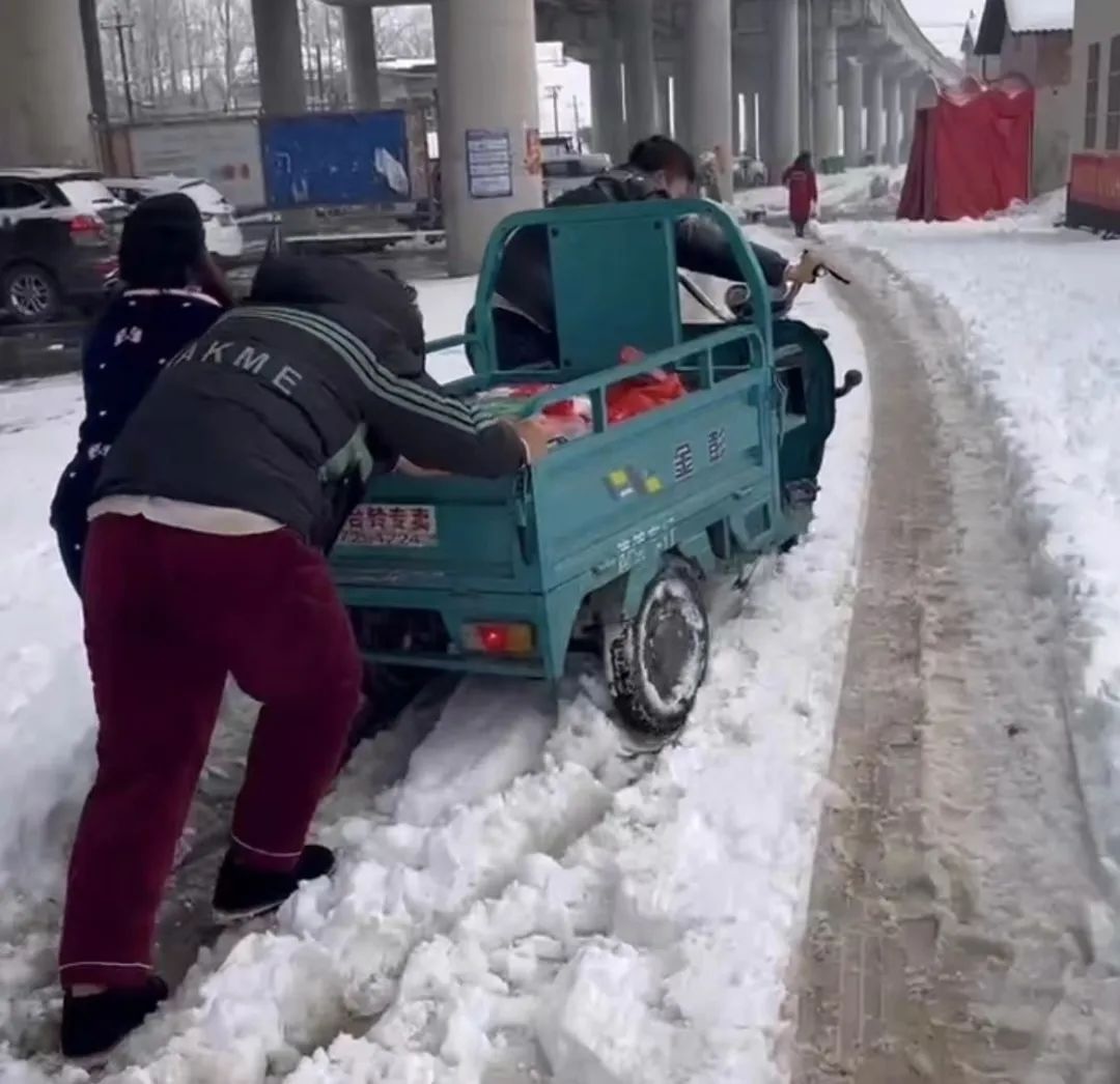 Three people push a blue three-wheeled motorized cart loaded with goods through heavy snow.