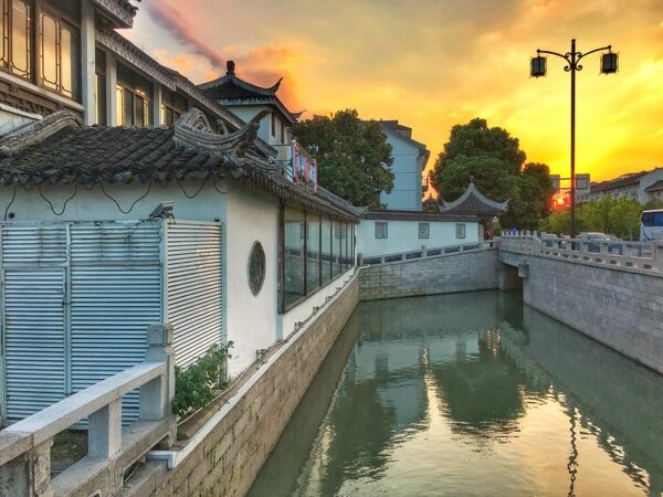 A brilliant yellow sunset is reflected in the water of a canal, which is lined with gray brick walls and charming traditional one- and two-story buildings with white walls and ornate black-tiled rooftops.