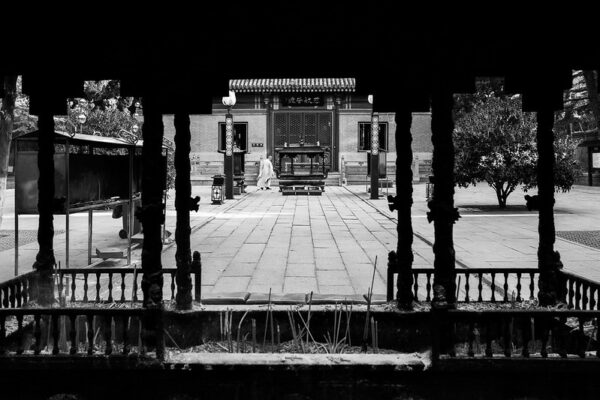 A study in perspective reveals the dark pillars of an overgrown pavilion in the foreground, and a long cobblestone path leading to the wooden door of a temple in the background.