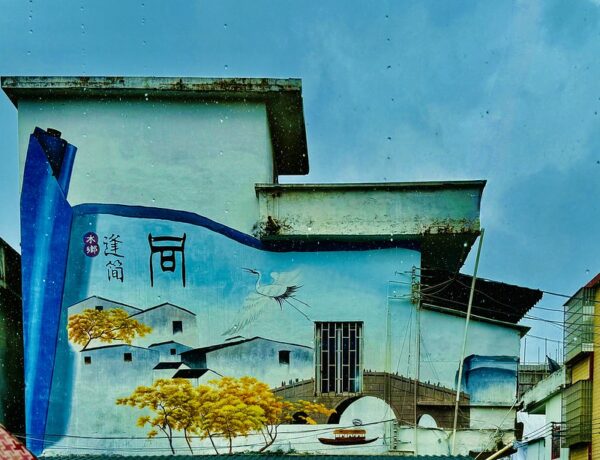 On the side of a house painted aquamarine, a mural in shades of pale blue, dark blue, white, and aquamarine vividly depicts the village of Fengjian with blue houses, yellow-leaved trees, a boat sailing under an arched stone bridge, and a bird soaring through a blue sky.