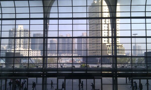 A view from within Shanghai’s high-speed railway station shows passengers walking to and fro and an enormous multi-story panel of windows framing some of Shanghai’s high-rise buildings.