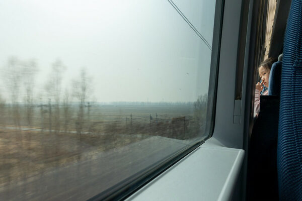 A little girl peeks between the seats of a modern high-speed train to look out a large glass window at the scenery rushing by: a gray sky, some leafless trees, and power lines.