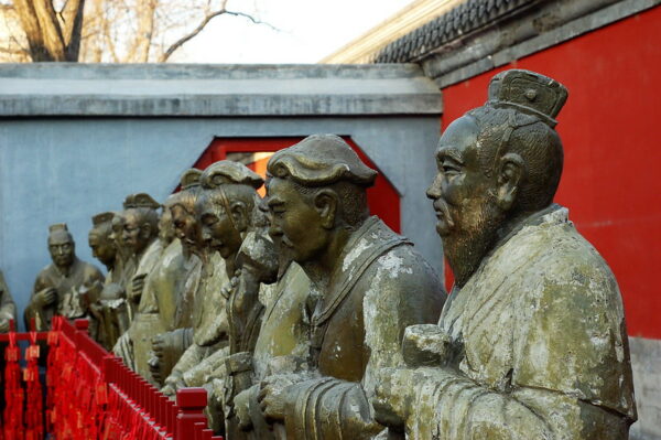 Against gray and deep vermillion walls topped with ceramic tile, a line of about a dozen stone statues stand, looking very somber and scholarly.