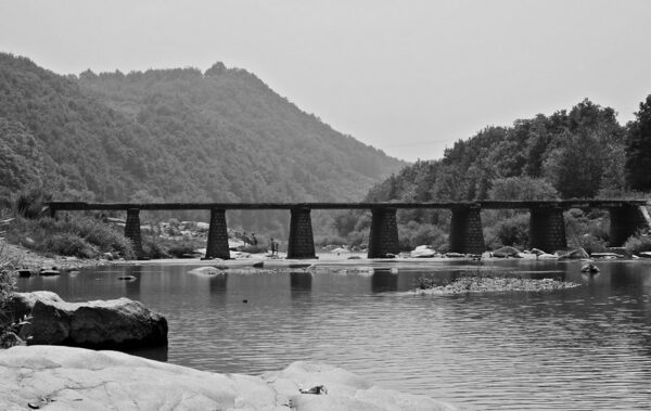 An old stone bridge supported by seven thick pillars spans a river. In the foreground are several large boulders, and in the background, some thickly forested hills.