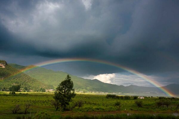An elongated rainbow stretches across the green fields of a valley ringed by low hills. The sky above the rainbow is ponderous with low, dark, thunderclouds.