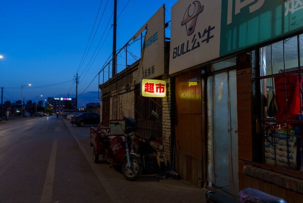 A road in Inner Mongolia at twilight is lined with above-ground power lines, street lamps, parked cars and motorized cycles, and several small shops, including a small “supermarket” marked by an illuminated red and yellow sign.