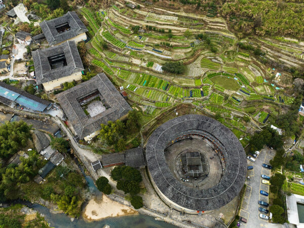 An aerial view of four enormous tiled-roofed tulou, a traditional architectural form of the Hakka people of southern China, surrounded by terraced fields in myriad shades of green. Three of the tulou are square, with large square courtyards at the center of each, and the fourth is round, with a vast circular courtyard at the center.