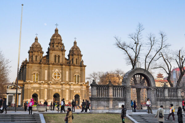 Dozens of people dressed in winter clothing walk, stand, and gaze at the main gate and brick facade of Wangfujing Cathedral (also known as the Church of St. Joseph) in Beijing. The Romanesque Revival-style cathedral is noted for its mixture of European and Chinese design features.