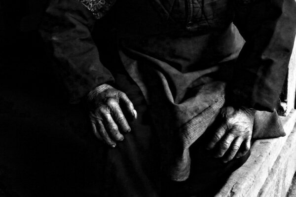 Close-up of the wrinkled, storied hands of an eighty-five-year-old rural resident of Tongnan, Chongqing, Sichuan province. The person is wearing what appears to be traditional, dark-colored clothing, possibly hand-dyed, with long sleeves.