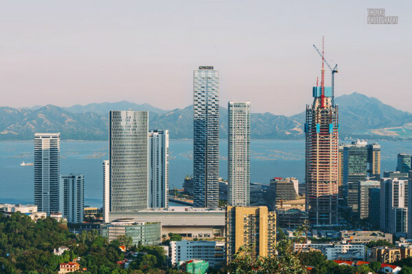 A stunning vista of Shekou’s forest of ultramodern skyscrapers, one under construction, with a vast expanse of water and some distant hills behind them.