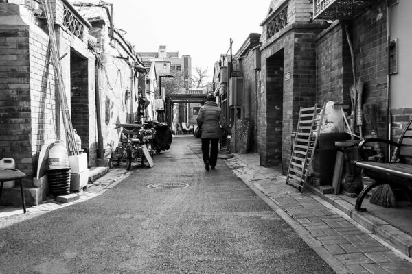 Seen from behind, a woman carrying a purse and wearing a warm hat and puffy jacket walks down a narrow alleyway lined with single-story brick dwellings and various bikes, mopeds, tools, brooms, and pieces of furniture.