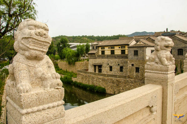 Two guardian lions adorning a light-colored stone bridge appear to be grinning. Nicely framed in the space between them is a two-storied, tile-roofed building of light-colored stone, with a stone wall separating it from what looks to be a narrow canal.