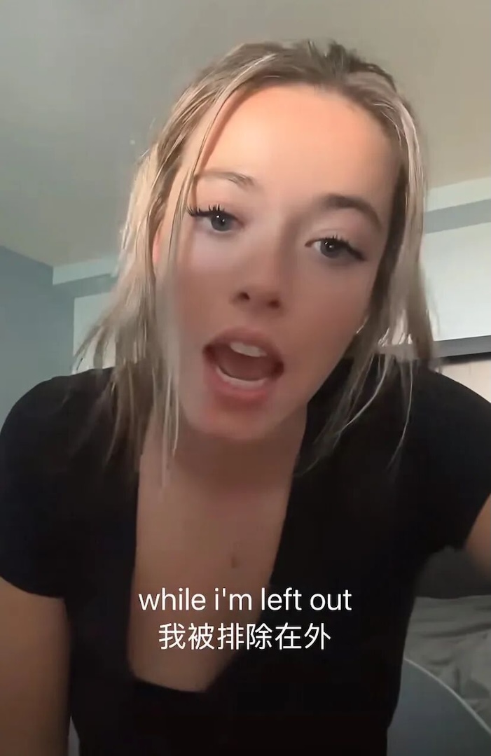 A social media post shows a young woman with blond hair and a black short-sleeved t-shirt speaking to the camera, with a bilingual Chinese and English caption that reads, “while i’m left out.”