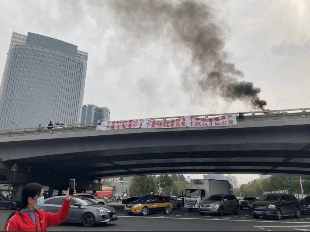 A column of smoke rises from a traffic overpass, on which a long banner hangs: “We want food, not COVID tests; reform, not Cultural Revolution. We want freedom, not lockdowns; elections, not rulers. We want dignity, not lies. Be citizens, not slaves.”
“Boycott classes. Boycott work. Depose the traitorous despot Xi Jinping.”