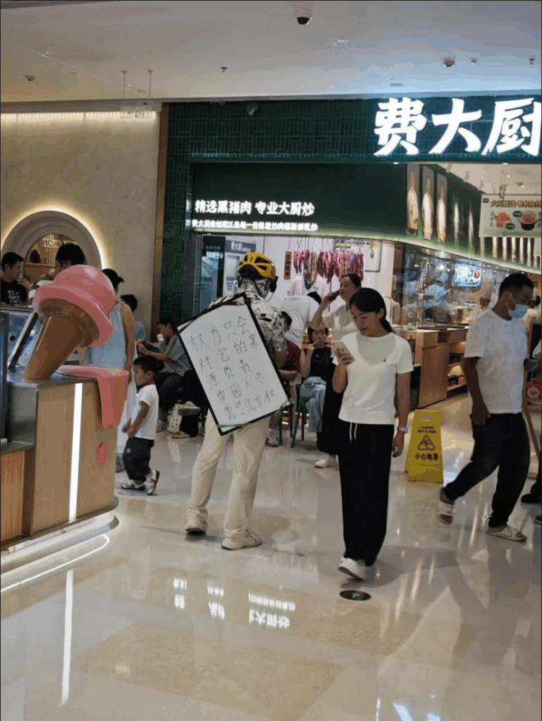 A man in a yellow bicycle helmet stands in a mall food court with a placard slung across his back: "Power is only accountable to its source. The Chinese people demand the right to vote."