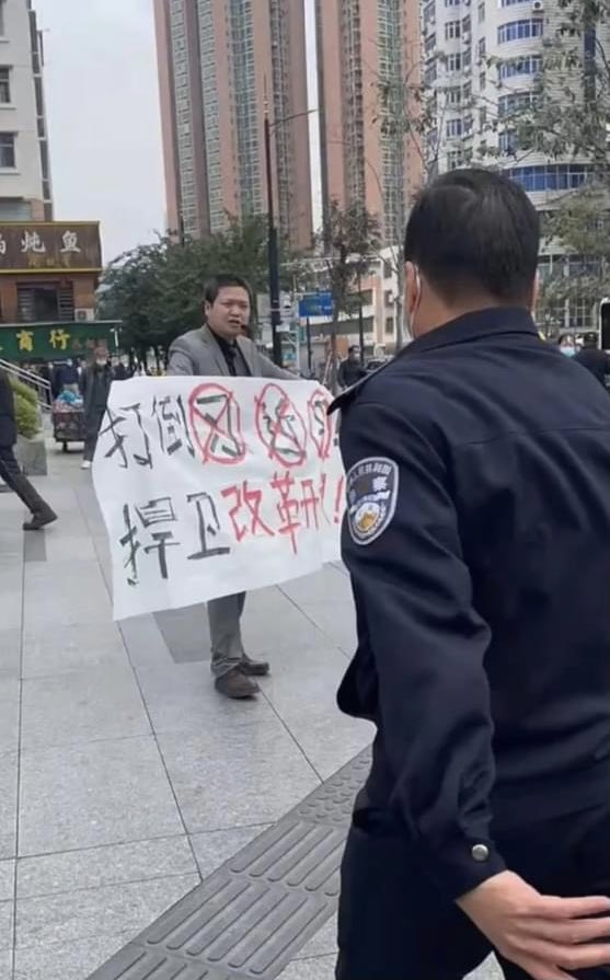 A man stands on the sidewalk, facing a police officer who has his back to the camera, and holding a banner that says "Down with Xi Jinping! Defend Reform and Opening!"