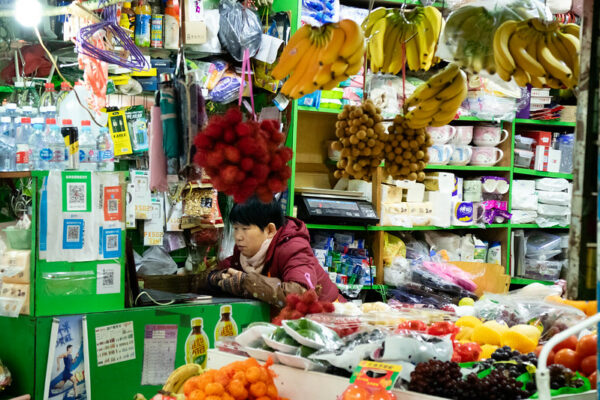 The proprietor of a small sundry goods shop gazes at a computer screen. She is dressed in a red padded jacket, gloves, and arm warmers. There is a counter heaped with fruits and vegetables, bunches of bananas and lychees hanging overhead, as well as some metal hangers, umbrellas, and various sundries. A wall on the side of the tiny shop has many cubbyholes stocked with daily necessities such as toilet paper, cups, tupperware, and kitchenware.
