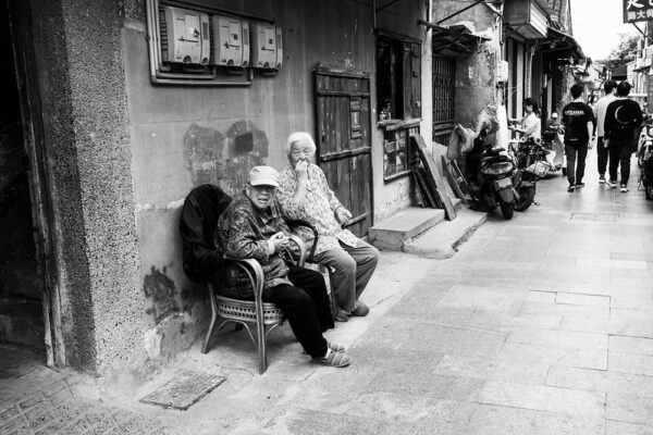 Dressed in slacks, colorful long-sleeved shirts, and comfortable slippers, two men who appear to be in their 70s or 80s sit beside each other in rattan chairs outside the door of a house and watch the passersby in a narrow, crowded alleyway.