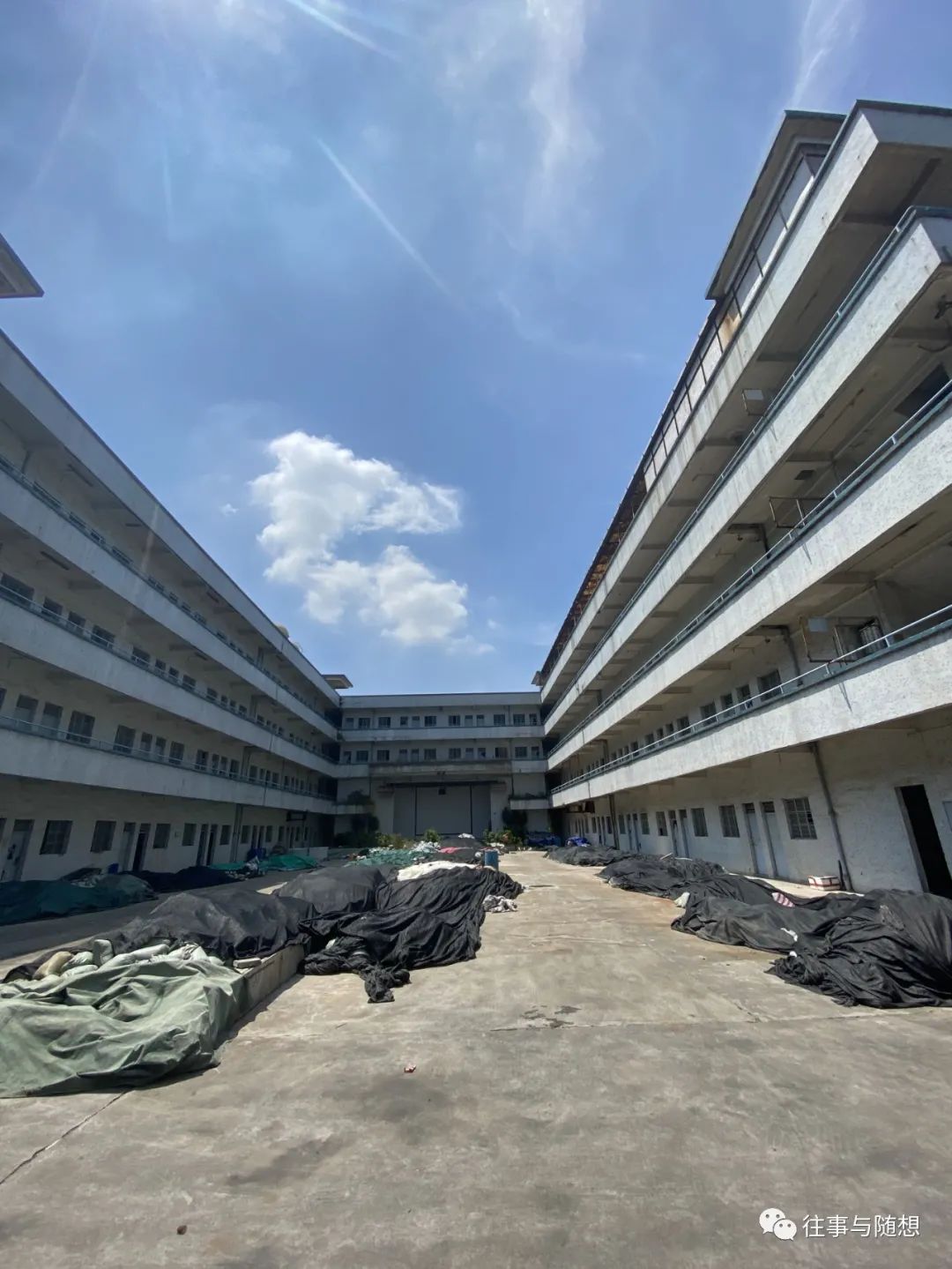 Under a bright blue sky, a rectangular courtyard surrounded by four-story concrete building. The courtyard is heaped with debris covered with green and black tarps.