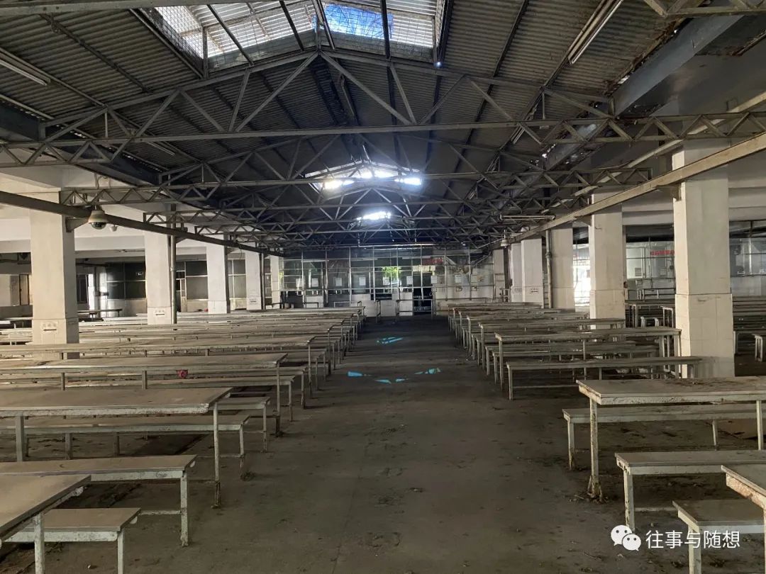 An abandoned dining hall with dozens of long table, concrete floors, concrete pillars and a corrugated metal roof with skylights.