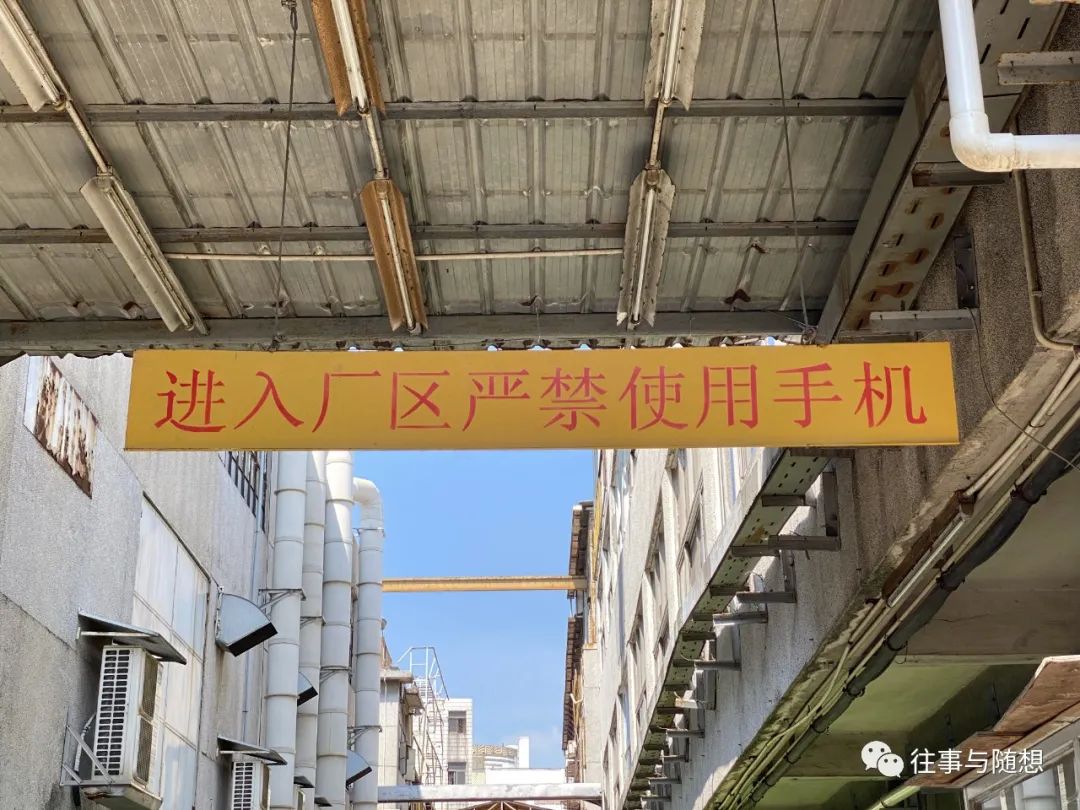 In a breezeway between two factory buildings, a large yellow overhead sign with red Chinese characters cautions that cell phone use is prohibited on factory premises.