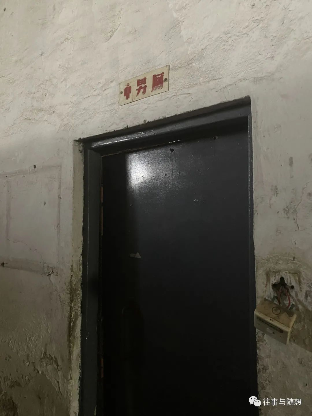 A black door in a very dirty white wall. A sign over the door reads "men's room" in red Chinese characters.