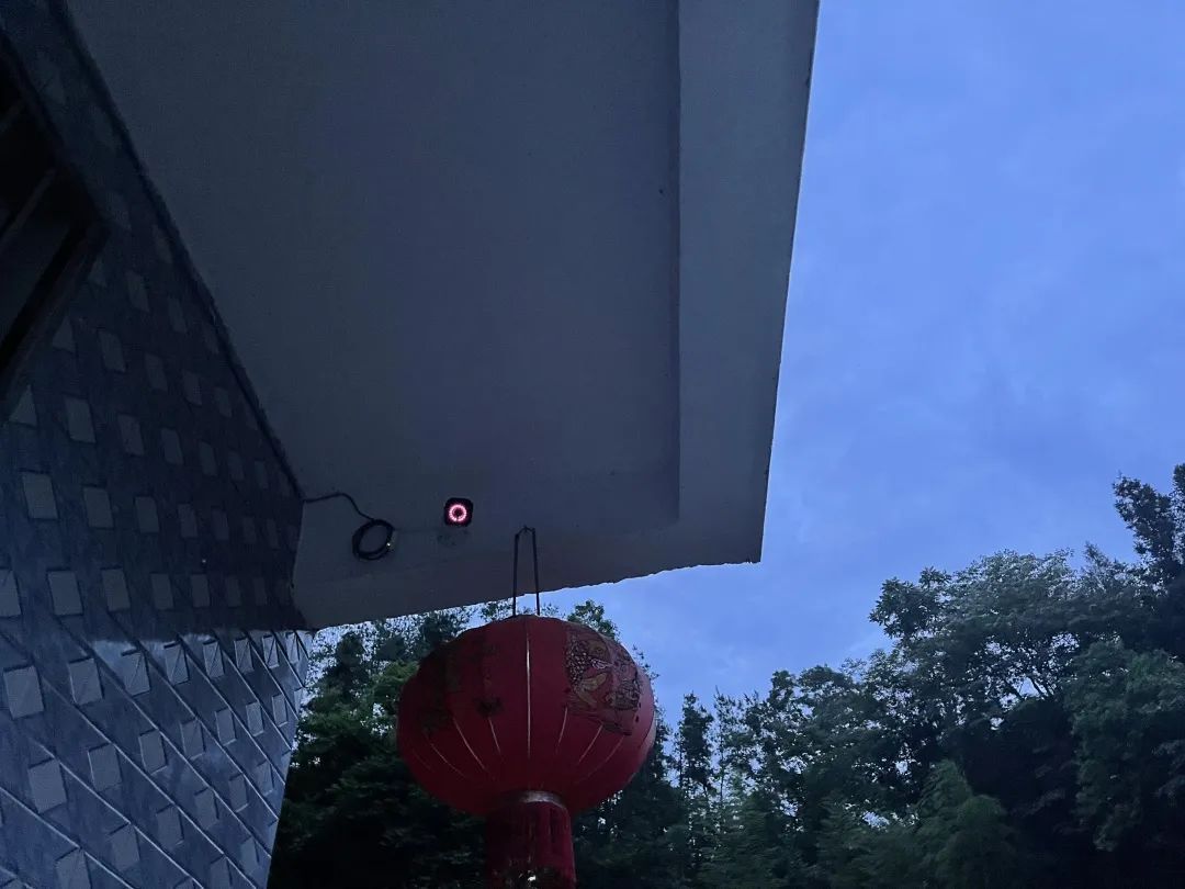A festive, traditional red lantern and a glowing surveillance camera adorn the eaves of a blue-tiled house.