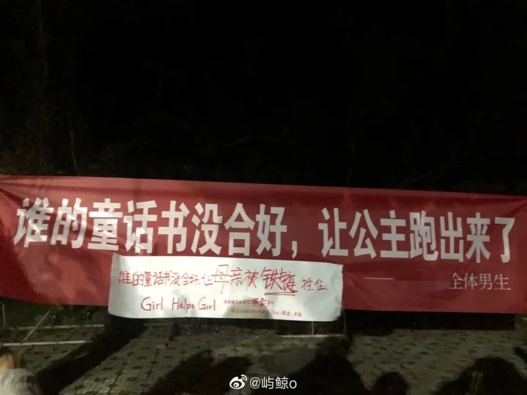 A red banner with white text sends a “fairytale” message, while a handwritten white poster with red text offers a rebuttal. Although it is night, the shadows of several people (likely those who affixed the poster to the banner) are visible on the pavement. 