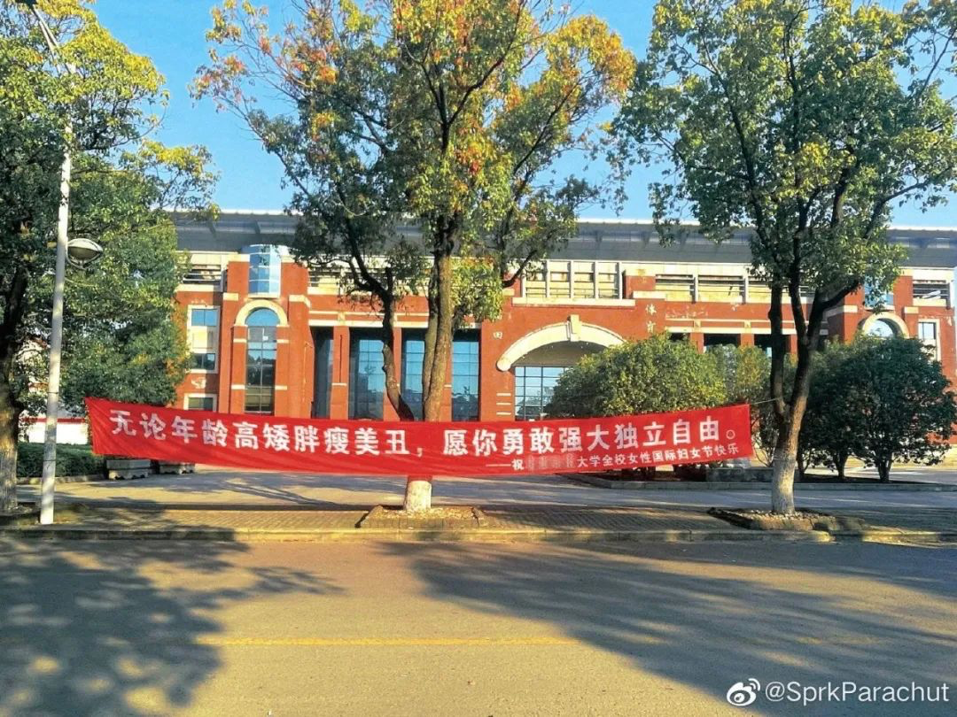 A red banner with white text is displayed along a campus road on a sunny day. 