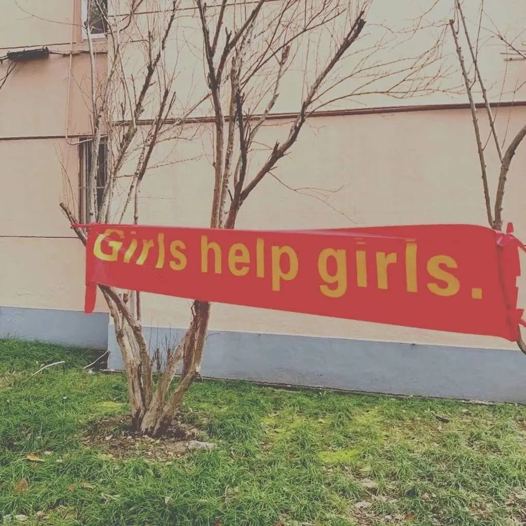 This red banner with yellow text, hung between two scrawny trees, displays a message in English: “Girls help girls.”