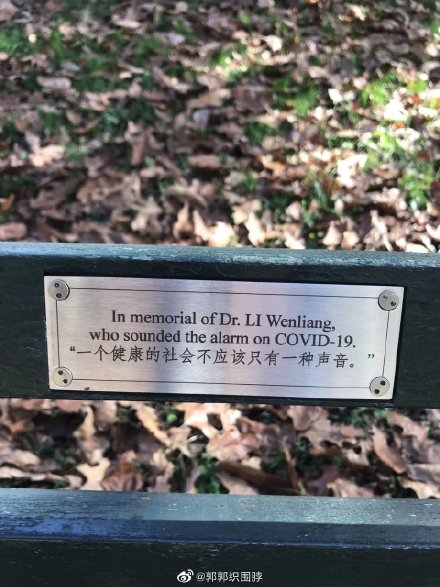 In a leafy park, a small metal sign on a green park bench reads: “In memorial of Dr. Li Wenliang, who sounded the alarm on COVID-19.”