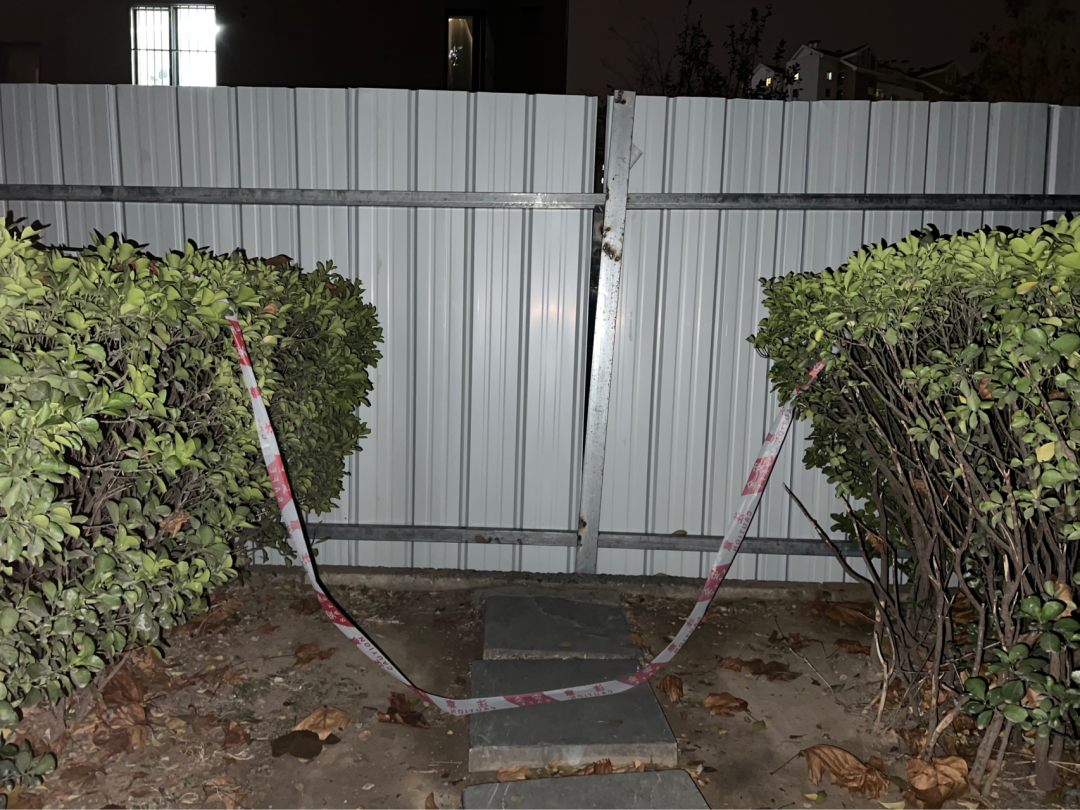 A sheet-metal fence, gray side facing inward, blocks a pedestrian path composed of large flat stones. A length of red and white caution tape has been draped over the path, and over the bushes on either side.