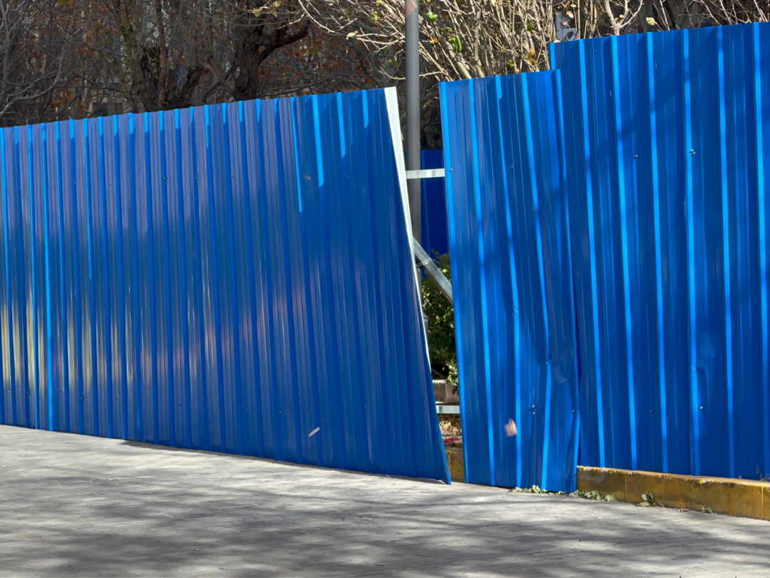 Missing panels create a large gap in a blue sheet-metal fence.