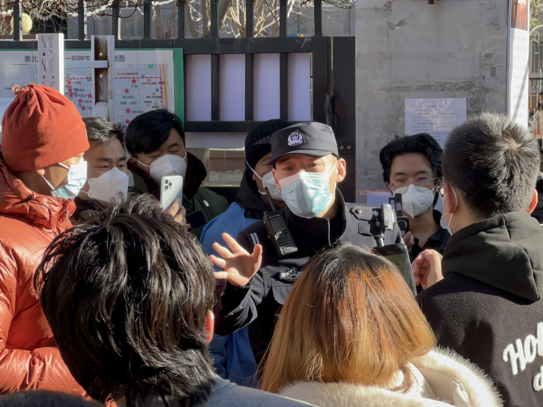A crowd of residents surround a male police officer, who appears to be conversing with them. Everyone is wearing heavy coats and surgical face masks, or N95 masks.