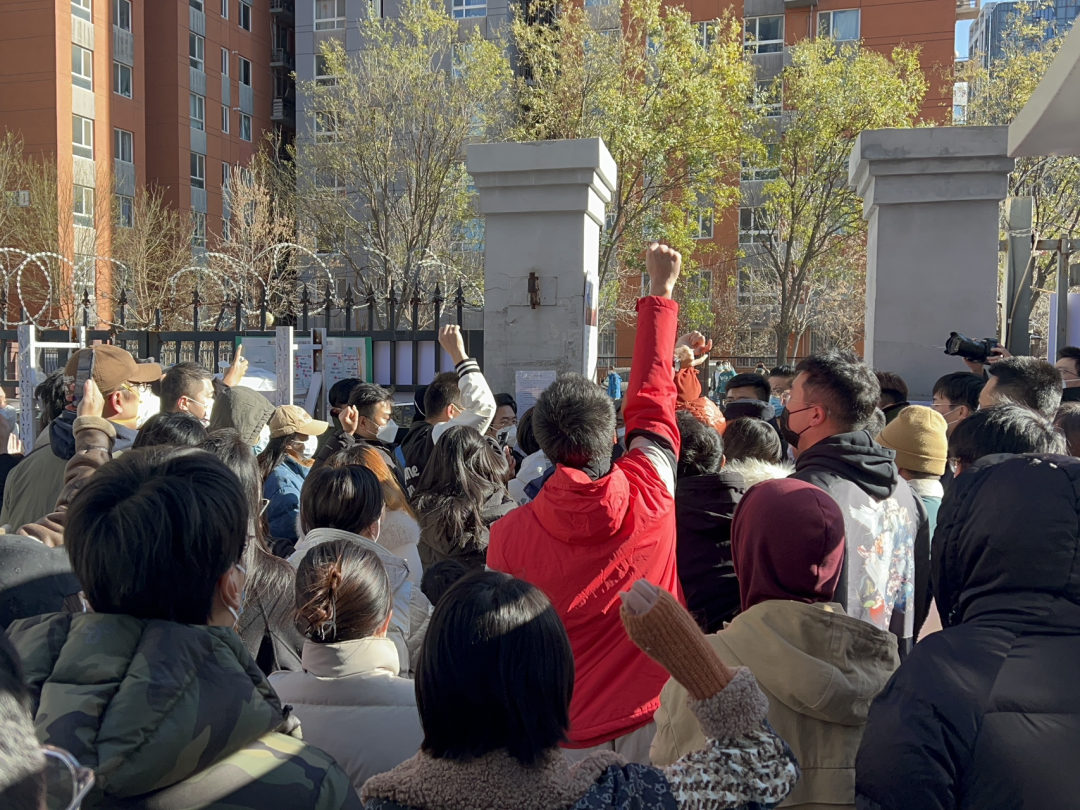 A crowd of residents stand near the gate and shout slogans. A few raise their fists into the air.