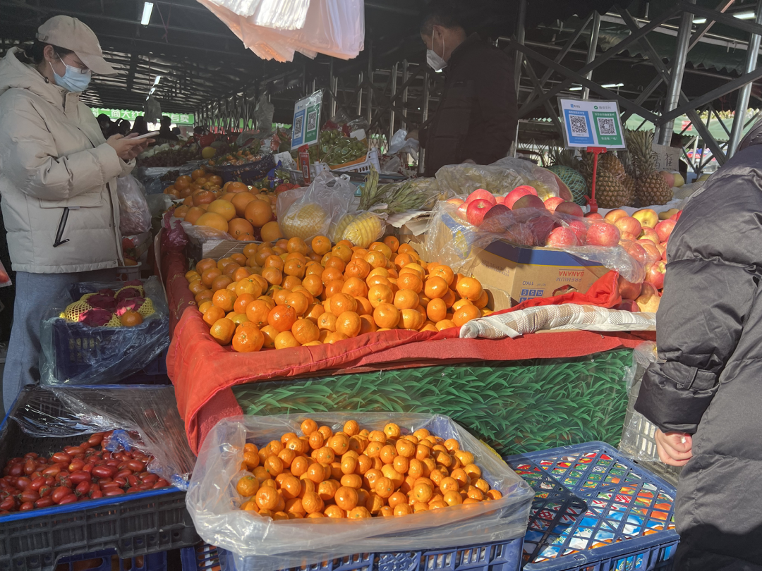 Several shoppers inspect the produce (apples, tangerines, and tomatoes) on display at a large fresh fruit-and-vegetable market.