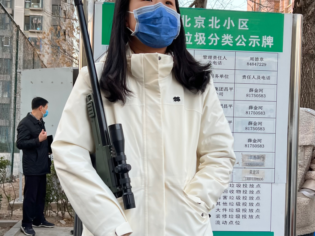 A young woman in a white jacket and blue surgical mask has a very realistic-looking toy rifle resting against her shoulder.