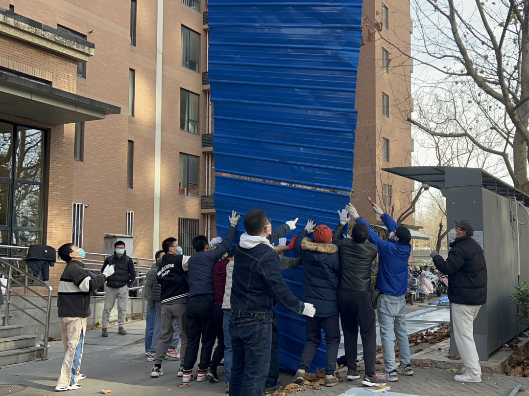 About a dozen residents help to hoist a large section of blue sheet-metal fencing into the air.