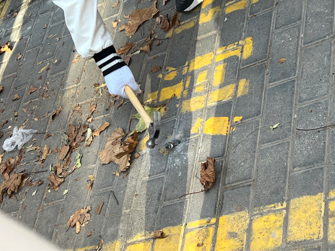A gloved hand grips a hammer above a large metal spike that has been flattened so that it doesn’t stick out too much from the pavement.