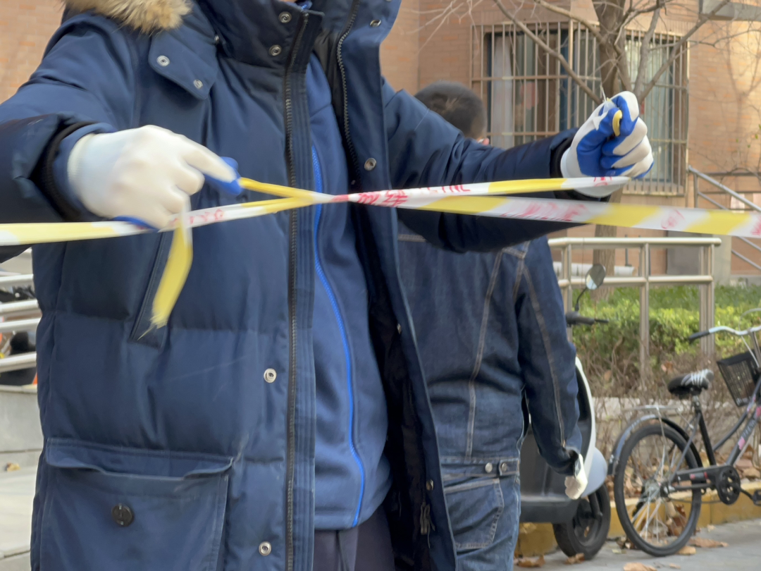 A man wearing white gloves, a navy fleece jacket, and a navy overcoat ties together two broken ends of yellow and white caution tape. His face is not visible.