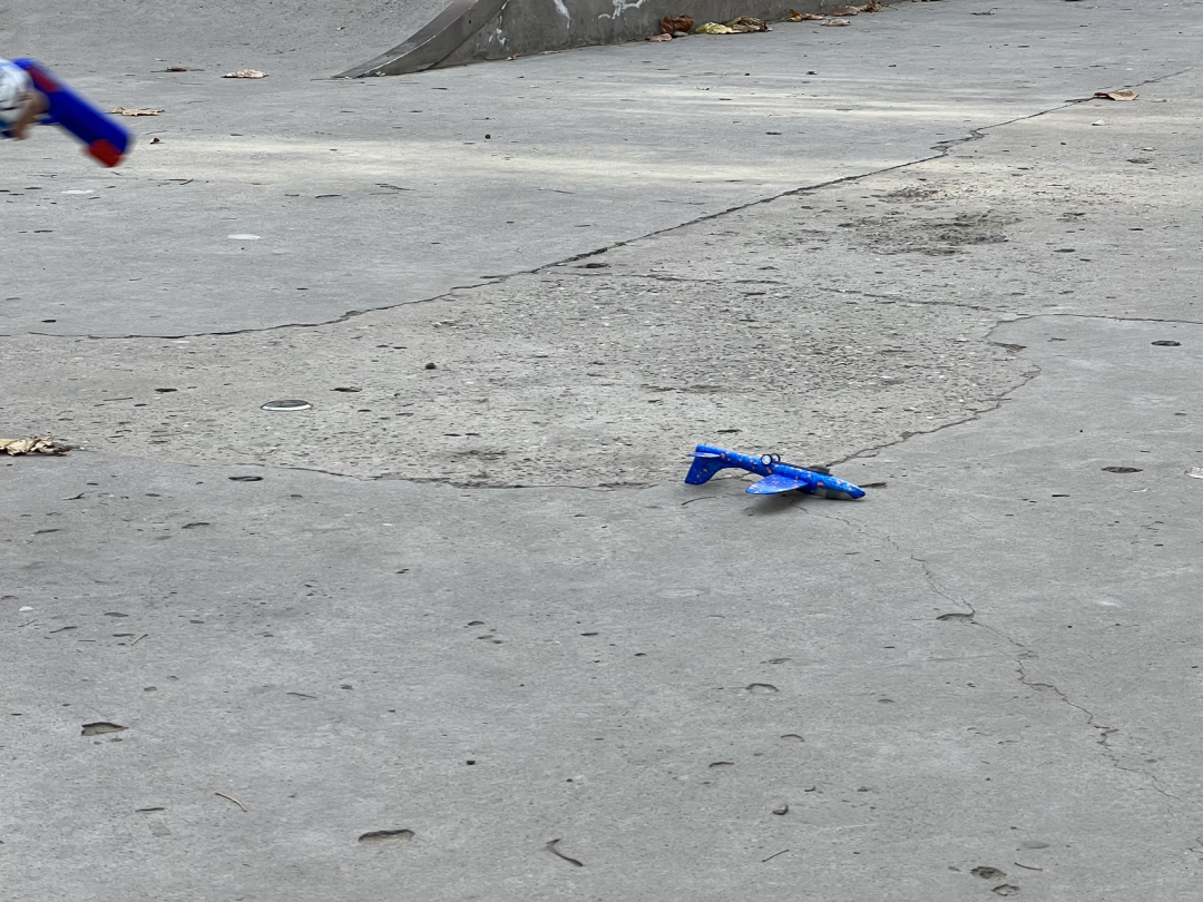 A blue plastic toy airplane lays on the pavement, looking rather the worse for wear. At the top left hand corner of the photo, just barely visible, a hand holds the blue plastic toy gun that the little airplane was launched from.