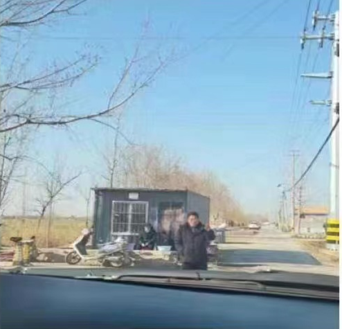 In this photo taken from behind the windshield of a car, a man dressed in black blocks the road ahead by standing directly in front of the car. Behind him, a guard wearing a blue surgical mask sits in front of a guardhouse made from a metal shipping container. 