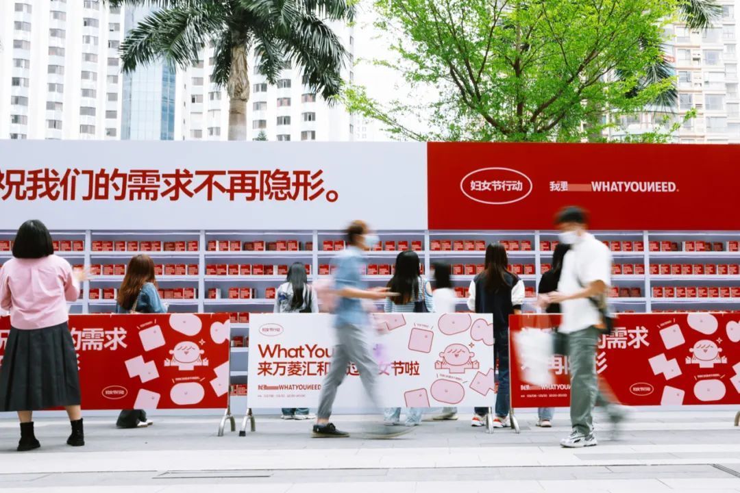 Eight women browse the shelves of an eye-catching red-and-white International Women’s Day pop-up store that has been set up along a busy sidewalk. In the foreground are the blurred figures of two male passersby who do not glance at the shelves.
