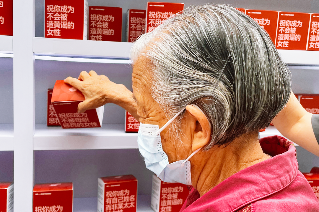 A gray-haired woman in a pink shirt and blue surgical mask selects a box that reads, “May you not be punished by motherhood.” Boxes on the shelves above and below feature other blessings, including “May you become a woman with a name of your own instead of simply ‘someone’s wife,’” and “May you not become the subject of sexualized rumors.”