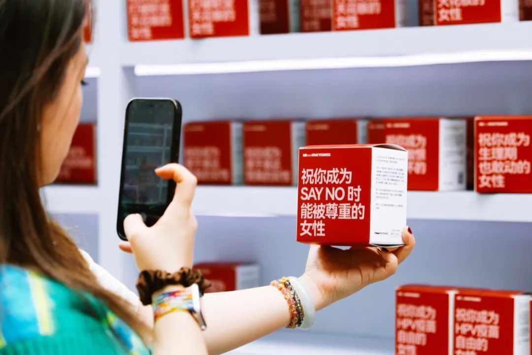 A young woman uses her cell phone to take a photo of a red-and-white box that reads, "May you be respected when you SAY NO."
